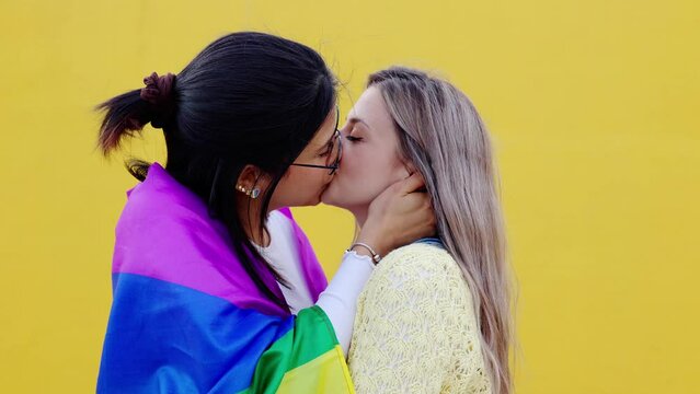 Two young generation z girls LGBTQ lesbian kissing and smiling at camera isolated on yellow background. Gay pride festival day concept.
