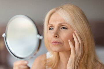 Upset senior woman checking wrinkles around her eyes, looking in mirror and touching face, making...