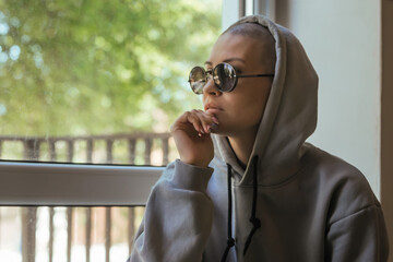 A beautiful young woman with a shaved head without hair sits and looks out the window, she is wearing dark glasses and a hoodie