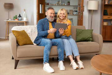 Cheerful Elderly Spouses Using Tablet Sitting On Couch At Home