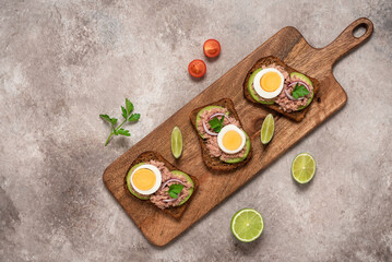 Obraz na płótnie Canvas Tuna toast. Open sandwich with tuna, rye bread, avocado and boiled egg on a wooden long board, brown grunge background. Top view, flat lay.