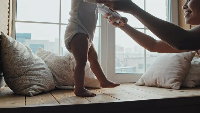 Barefoot baby slowly walking along wooden window sill, supported by joyous mother