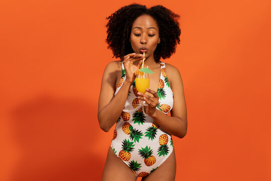 Happy young black woman in colorful one-piece swimsuit drinking cocktail