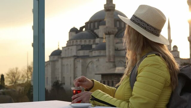 Happy attractive woman tourist in hat, drinks traditional turkish tea (armudas) against the background of Suleymaniye mosque, Istanbul, Turkey. Religion and travel concept.