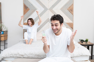 Obraz premium Happy man sitting on the bed, holding tablets and demonstrating his happiness after solving his erectile dysfunction problem