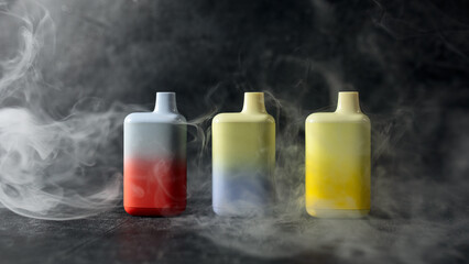 A set of multicolored disposable electronic cigarettes on a dark background stand on a table with smoke around. The concept of modern smoking, vaping and nicotine