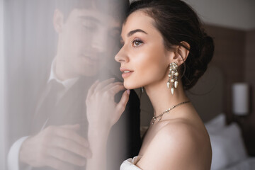 stunning bride in elegant jewelry and wedding dress hugging shoulder of groom in classic formal wear while standing together behind white tulle in modern hotel room after ceremony