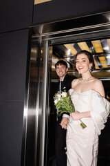 pretty bride in white dress holding bridal bouquet with flowers and hand of cheerful groom in suit walking out of elevator in hotel, happy newlyweds © LIGHTFIELD STUDIOS