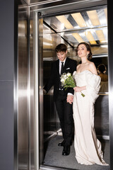 full length of pretty bride in white dress holding bridal bouquet with flowers and hand of cheerful groom in suit standing in elevator in hotel, happy newlyweds © LIGHTFIELD STUDIOS