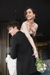 good looking groom in black formal wear lifting cheerful bride with makeup in white wedding dress holding bridal bouquet of flowers while standing in hotel lobby