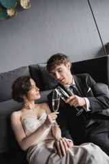 happy newlyweds, bride in white wedding dress and groom in black suit holding glasses of champagne while clinking and celebrating their marriage after wedding in hotel room