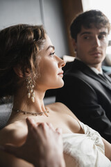 Naklejka premium elegant bride in white wedding dress and luxurious jewelry looking away near handsome groom in suit looking at her on blurred background of hotel room