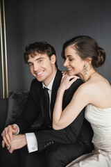 portrait of charming bride in white wedding dress smiling near handsome groom in black suit looking at camera while sitting in hotel room, happy newlyweds