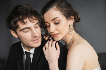 portrait of beautiful young bride in luxurious earrings with pearls and necklace sitting with closed eyes and hugging happy groom in black suit in hotel room