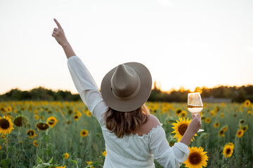 Portrait of beautiful young woman 33 years old in hat in sunflower field at sunset holds glass of white wine in her hand. Happy model in white dress on a summer evening in nature. Warm.