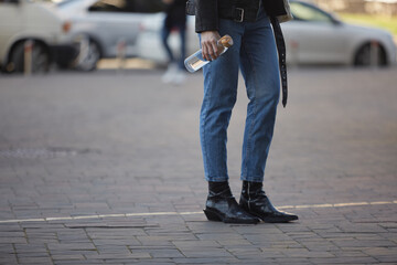 Young woman walking with a glass water bottle outdoor. Unrecognizable female person wearing blue skinny jeans and stylish leather boots on a walk in the city center
