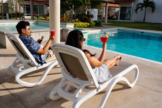 A young latin couple relaxing in a sun bed by the poolside and drinking fresh delicious watermelon drinks