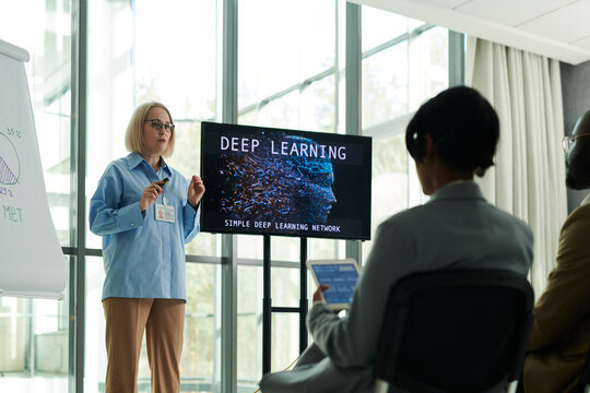 Confident blond mature woman in smart casualwear making presentation of new educational project on interactive board to her learners