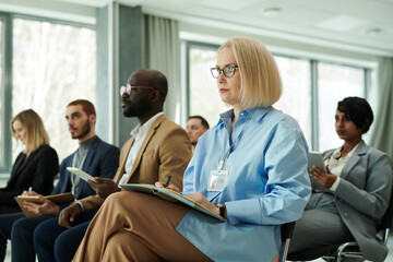 Blond serious businesswoman with open notebook on her knees looking at lecturer or speaker while...
