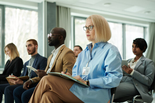Blond serious businesswoman with open notebook on her knees looking at lecturer or speaker while listening to report and making notes