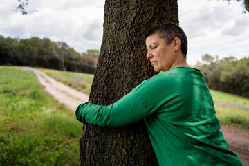 Non-binary woman with very short hair hugging a large oak tree in a field, green T-shirt and hair...