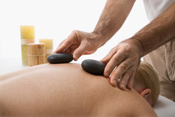 Caucasian middle-aged male massage therapist placing hot stones on back of Caucasian middle-aged woman lying on massage table.