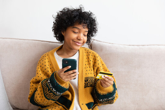 African american woman shopping online holding smartphone paying with gold credit card. Girl sitting at home buying on Internet enter credit card details. Online shopping ecommerce delivery service