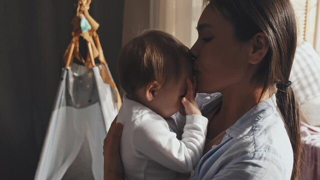 Medium close up of mom holding baby, kissing her in forehead with love and care at home
