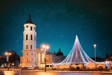 Vilnius, Lithuania. Amazing Bold Bright Blue Starry Sky Gradient Above Christmas Tree On Background Bell Tower Belfry Of Vilnius Cathedral At Square In Evening New Year Christmas Xmas Illuminations. - obrazy, fototapety, plakaty