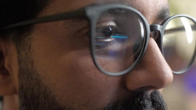 Close-up of bearded multiethnic programmer with brown eyes looking at monitor. Computer screen light reflecting from glasses. Handsome worker programming code. Creating software for websites or games.