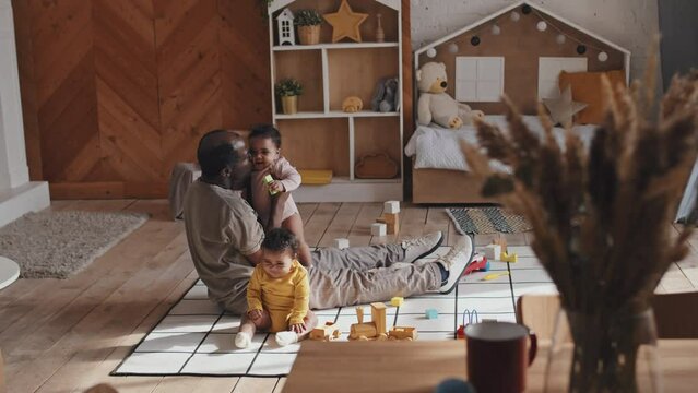 Full shot of loving African American father sitting on floor in nursery room, holding baby daughter on arms and kissing as her twin sister playing beside during day at home