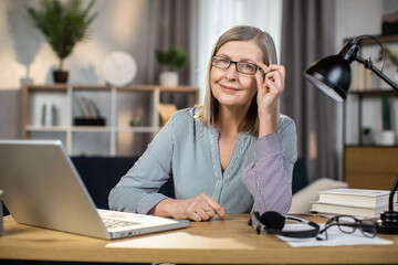 Portrait of happy mature lady in everyday clothes posing with laptop on writing desk in cozy workplace indoors. Cheerful caucasian female in spectacles using remote office for managing business.