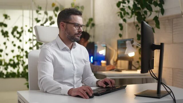 Good-looking Caucasian financial advisor using keyboard and with mouse scrolling web site. Attractive man working on computer in modern business center. Banking commerce. Economic development.