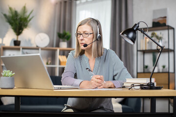 Middle-aged elegant woman using headset with microphone while enjoying home comfort during remote work. Smiling caucasian freelancer in eyeglasses holding online videoconference and making notes.
