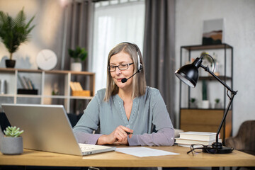 Middle-aged elegant woman using headset with microphone while enjoying home comfort during remote work. Smiling caucasian freelancer in eyeglasses holding online videoconference and making notes.