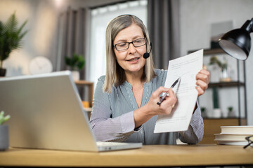 Focus on elderly lady in formal wear and glasses showing documents while sitting with headset at...