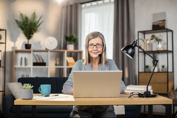 Portrait of smiling female worker in hands-free headset sitting behind laptop at office desk in...
