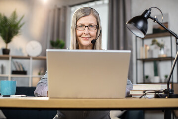 Attractive lady in early 60s with wireless headset smiling warmly while staying in home office with modern laptop. Confident freelancer enjoying benefits of telework using electronic devices.