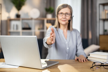 Focus on female arm stretched out for handshake by attractive woman of mature age spending working hours in remote office. Professional manager in wireless headset welcoming colleagues on meeting.