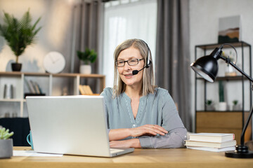 Portrait of smiling female worker in hands-free headset sitting behind laptop at office desk in...