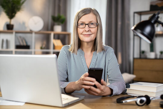 Mindful senior woman in spectacles looking at phone screen while operating modern laptop at office desk in home interior. Efficient freelancer checking meetings schedule via mobile application. - Powered by Adobe