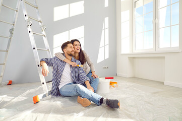 Happy young loving couple planning home repair while sitting on floor in living room. Cheerful wife hugging her husband in freshly painted room. Home repair and decoration concept