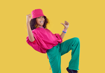 Little kid in trendy clothes. Happy child having fun in fashion studio. Afro American girl wearing fuschia bucket hat, loose oversized top and green jeans dancing isolated on solid yellow background - obrazy, fototapety, plakaty
