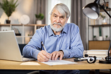 Confident caucasian man of mature age taking notes while utilizing portable computer in remote work from home. Skillful freelancer in business casual clothes writing initial report of project indoors.