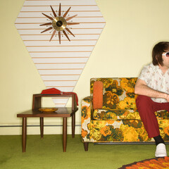 Caucasian mid-adult man sitting on colorful retro sofa wearing sunglasses in room with vintage decor.