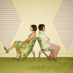 Caucasian mid-adult couple wearing vintage clothing sitting back to back in green vinyl chairs with...
