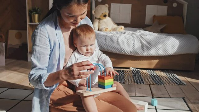 Cropped front medium shot of dark-haired casually dressed mom sitting on floor in sunlit nursery, holding baby, playing together educational game with multi-coloured bricks. Morning