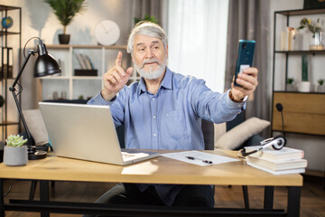 Smiling man about 65 posing with pointing finger in front of smartphone camera while taking break from work in home office. Mature freelancer taking selfie of comfortable lifestyle image indoors.