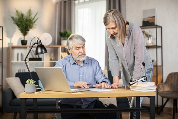 Attractive female standing with pen in hand while senior male looking through paper document on desk near laptop. Experienced entrepreneurs correcting written contract together in home office interior
