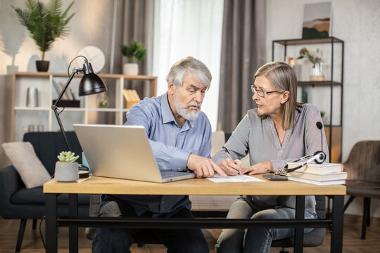 Middle-aged lady in glasses putting down information while senior male finding data on laptop in creative office. Efficient professionals drafting written agreement while doing distant work from home. - Powered by Adobe